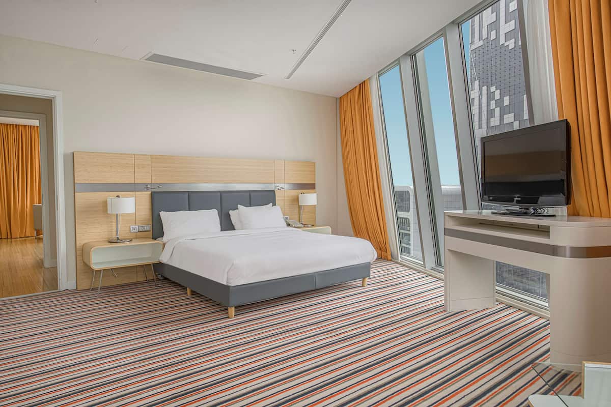 A modern hotel room with a large bed, a desk with a flat-screen TV, and a city view.