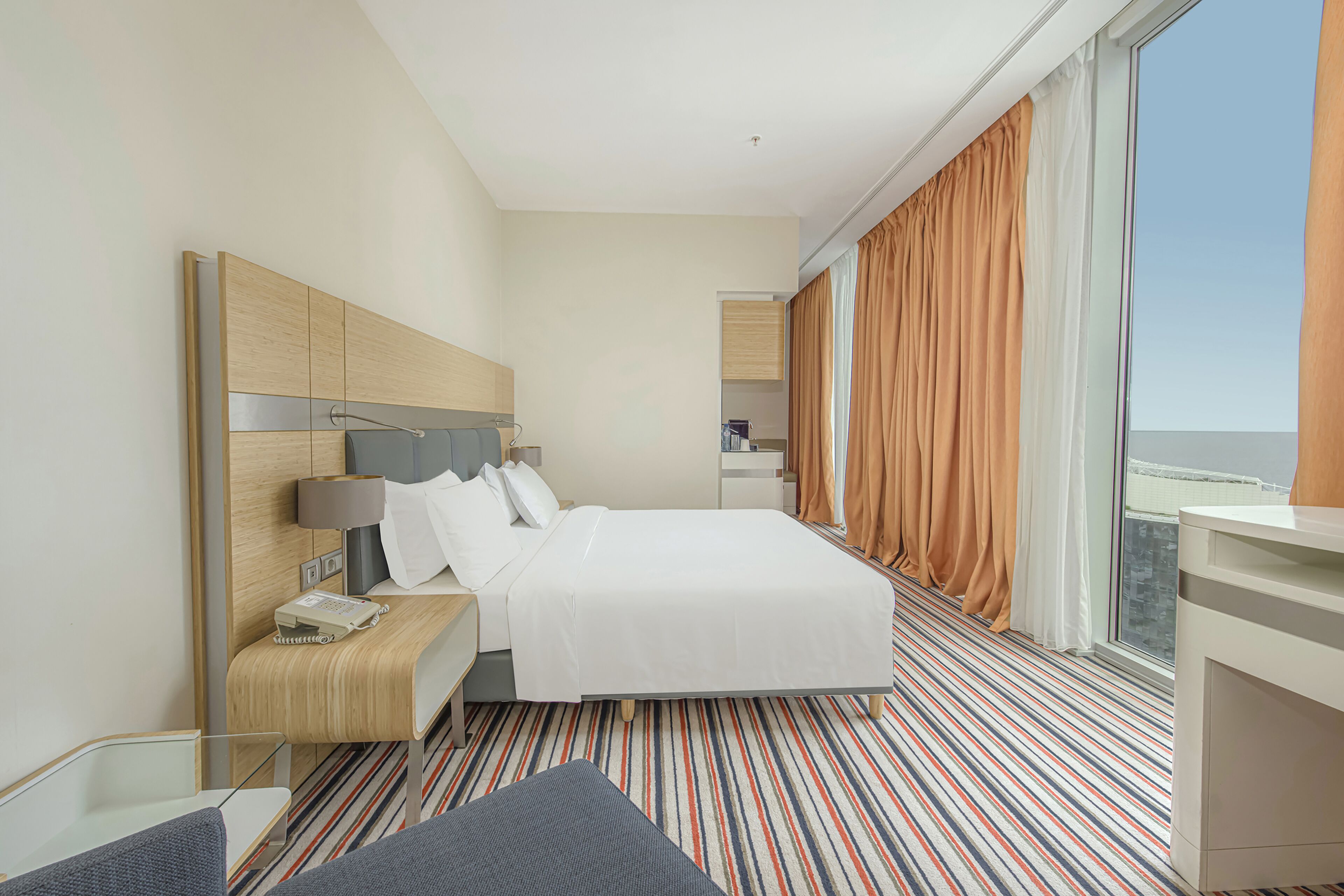 A hotel room with a large bed, a desk, a chair, and a view of the city through large windows.