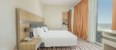 A hotel room with a large bed, a desk, a chair, and a view of the city through large windows.