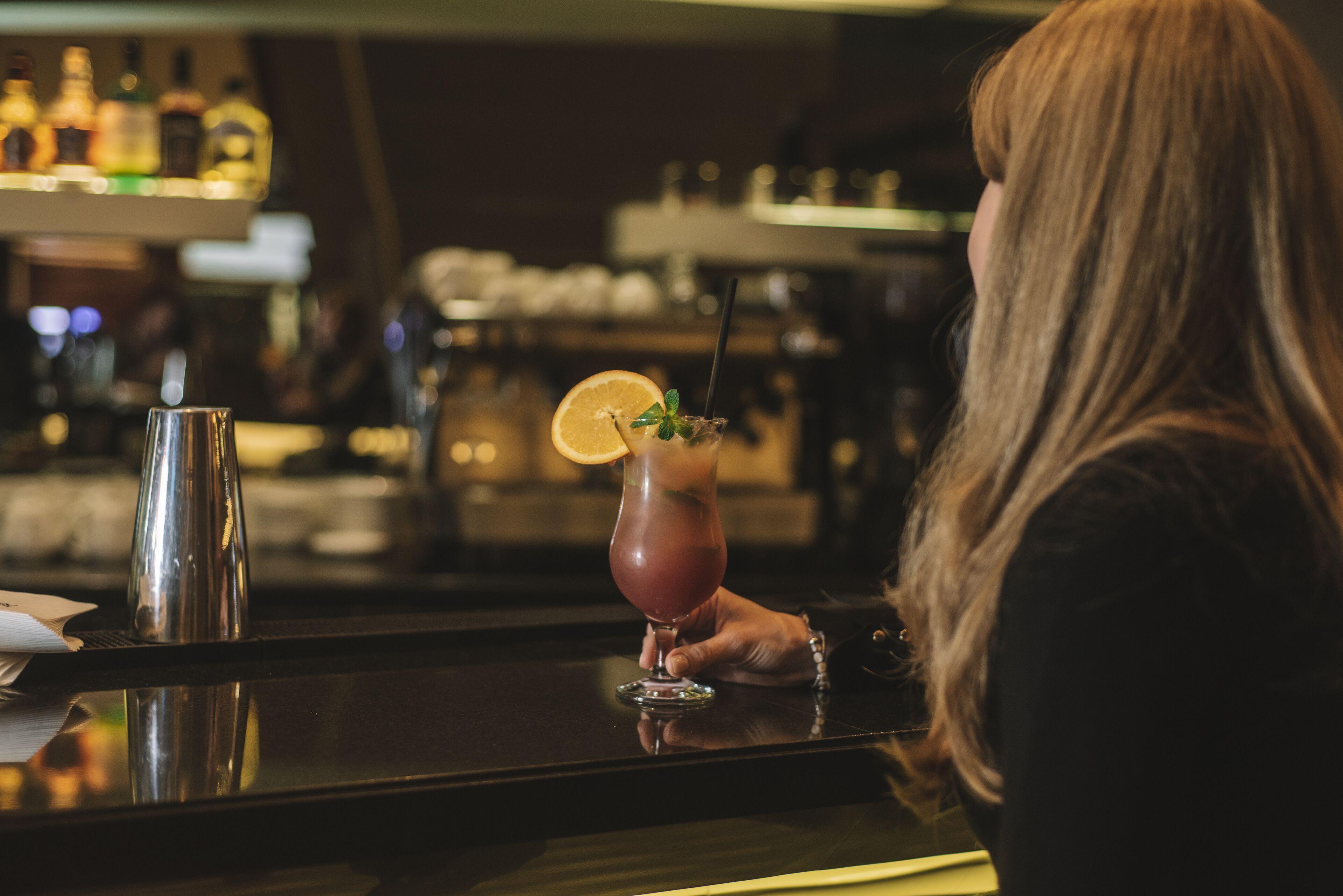 A bar setting with a person holding a drink, a bottle, and a glass on a reflective surface.