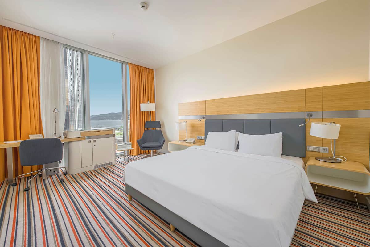 A hotel room with a large bed, a desk with a chair, a small table with a lamp, and a view of the city through large windows.