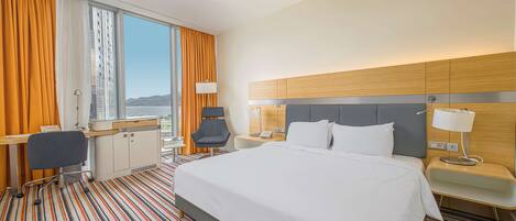 Una habitación de hotel con una cama grande, un escritorio con una silla, una mesita de noche con una lámpara y vistas a la ciudad a través de amplios ventanales.