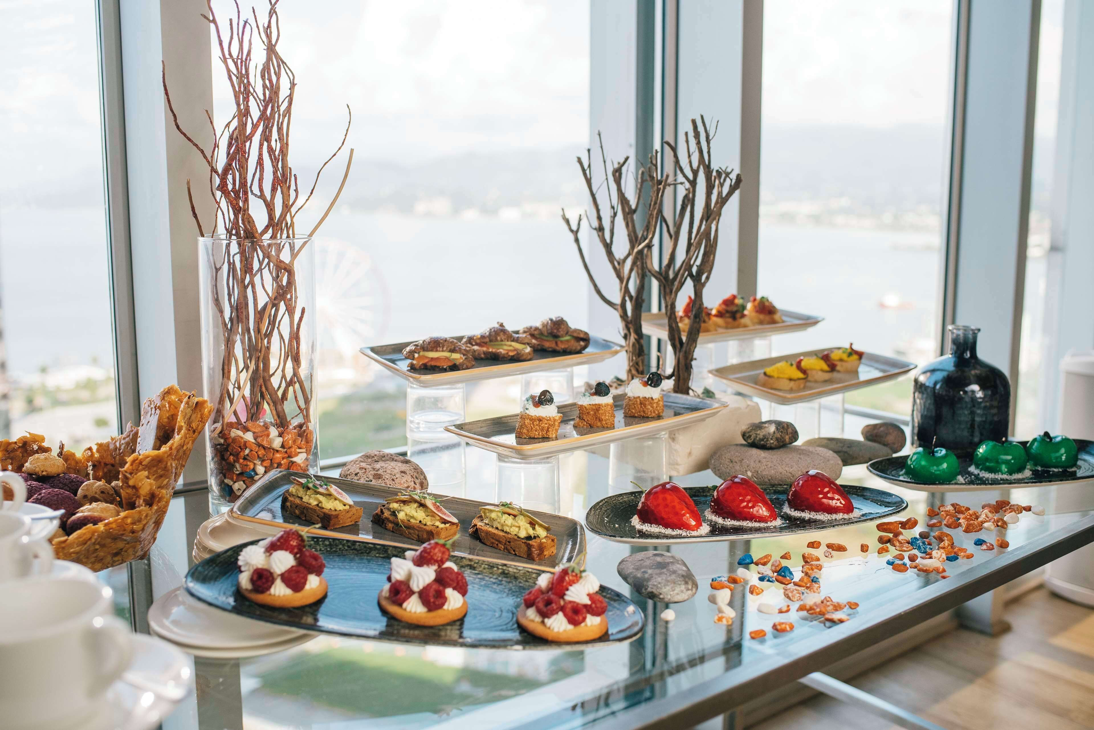 A buffet table with various desserts and snacks, set against a large window with a view of a cityscape.