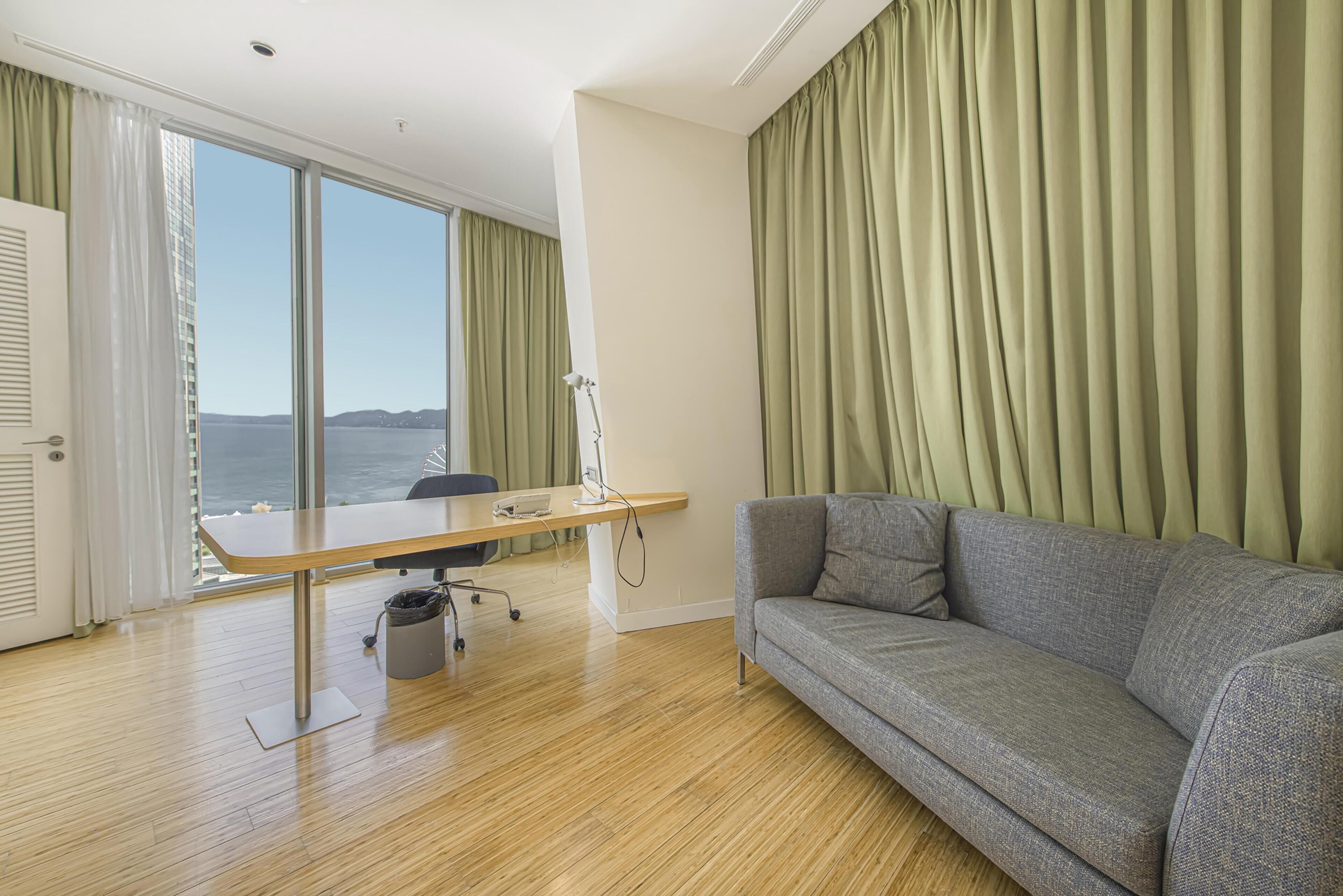 A room with a desk, a grey sofa, a view of the sea, and a window with curtains.