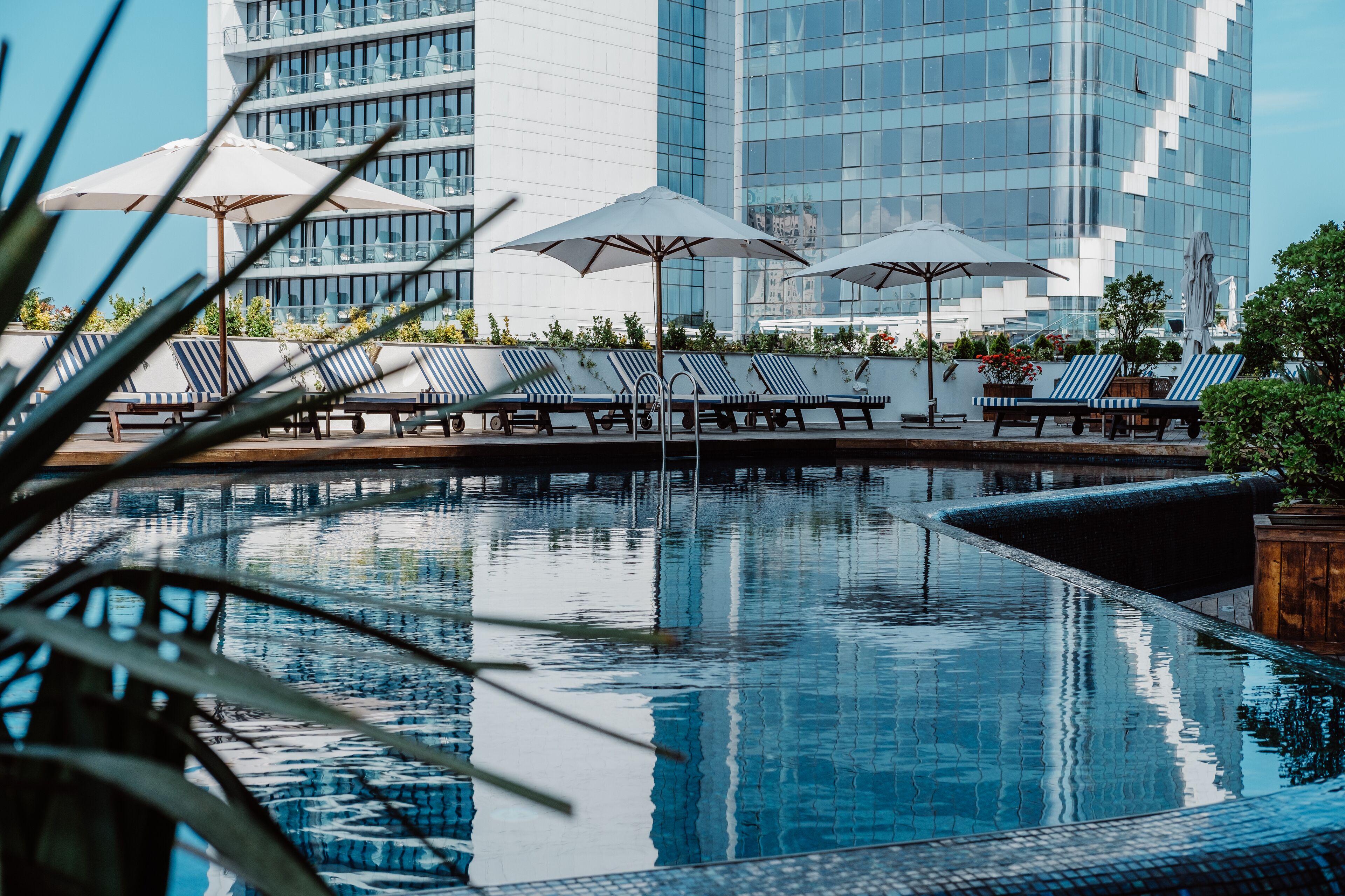 Un espace piscine agrémenté de chaises longues et de parasols, entouré d’un bâtiment moderne aux fenêtres en verre réfléchissant.