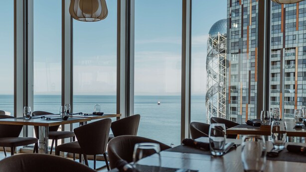 Ein Restaurant mit gedeckten Tischen, großen Fenstern mit Meerblick und einem modernen Gebäude mit Glasfassade im Hintergrund.