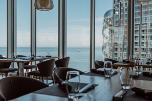 A restaurant with tables set for dining, large windows offering a view of the sea, and a modern building with a glass facade in the background. - Radisson Blu Hotel Batumi (Batumi)