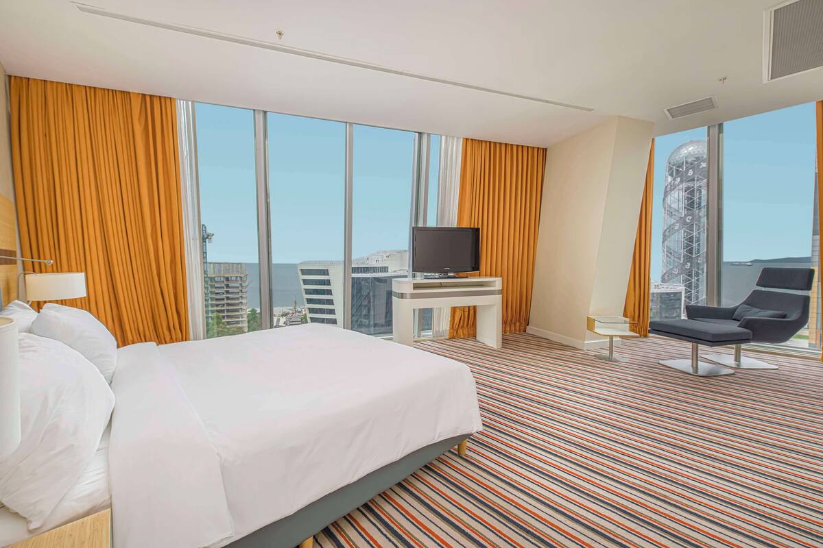 A hotel room with a large bed, a TV on a stand, a lounge chair, and large windows offering a city view.