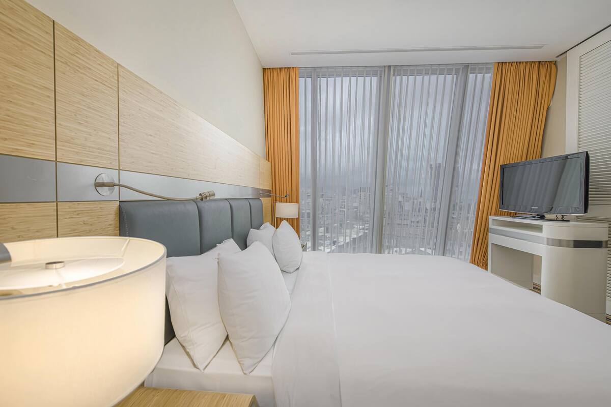 A hotel room with a bed, a TV, a desk, and a view of the city through sheer curtains.
