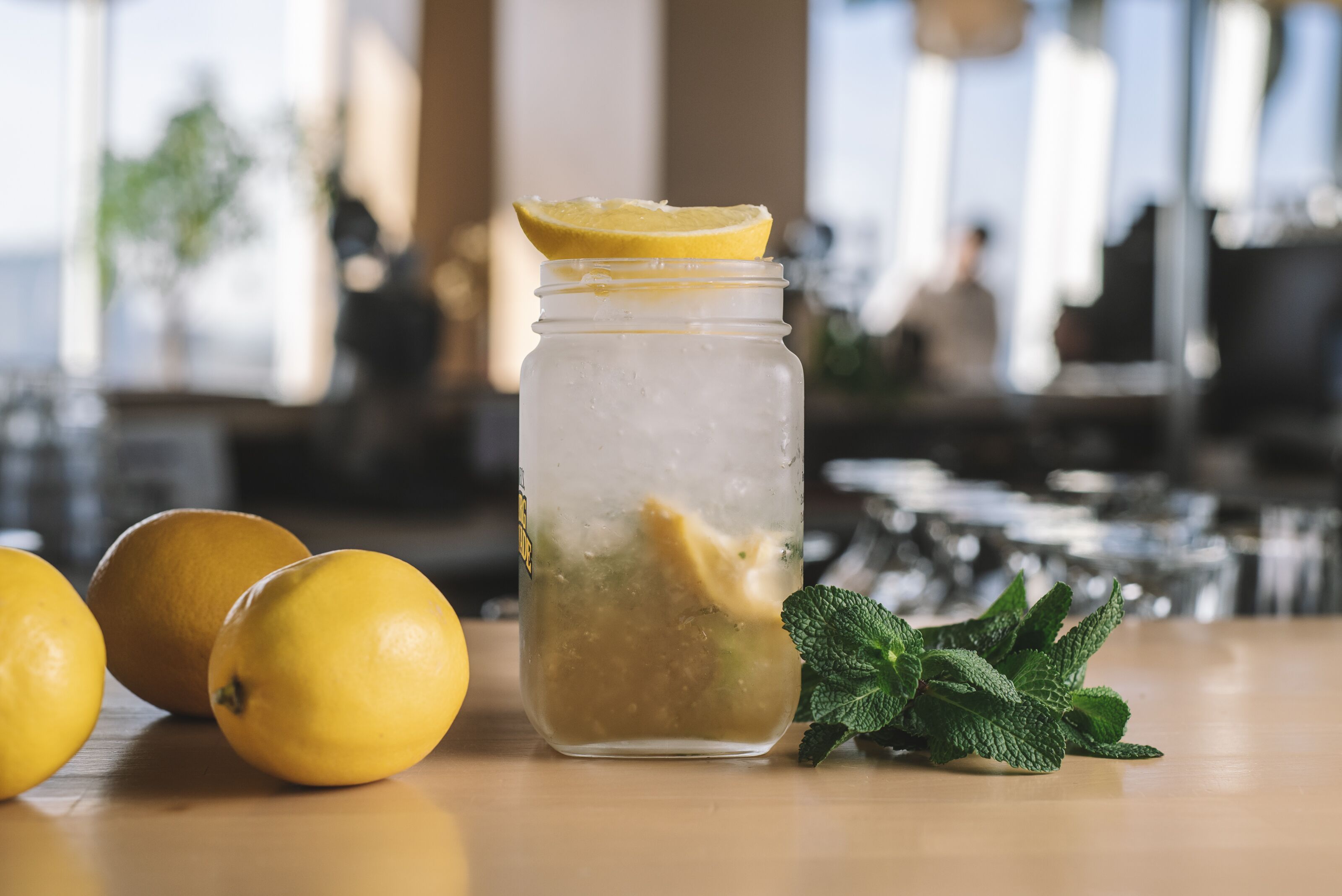 Un tarro de vidrio transparente con una rodaja de limón encima, lleno de un líquido claro, acompañado de limones enteros y hojas de menta sobre una superficie de madera.