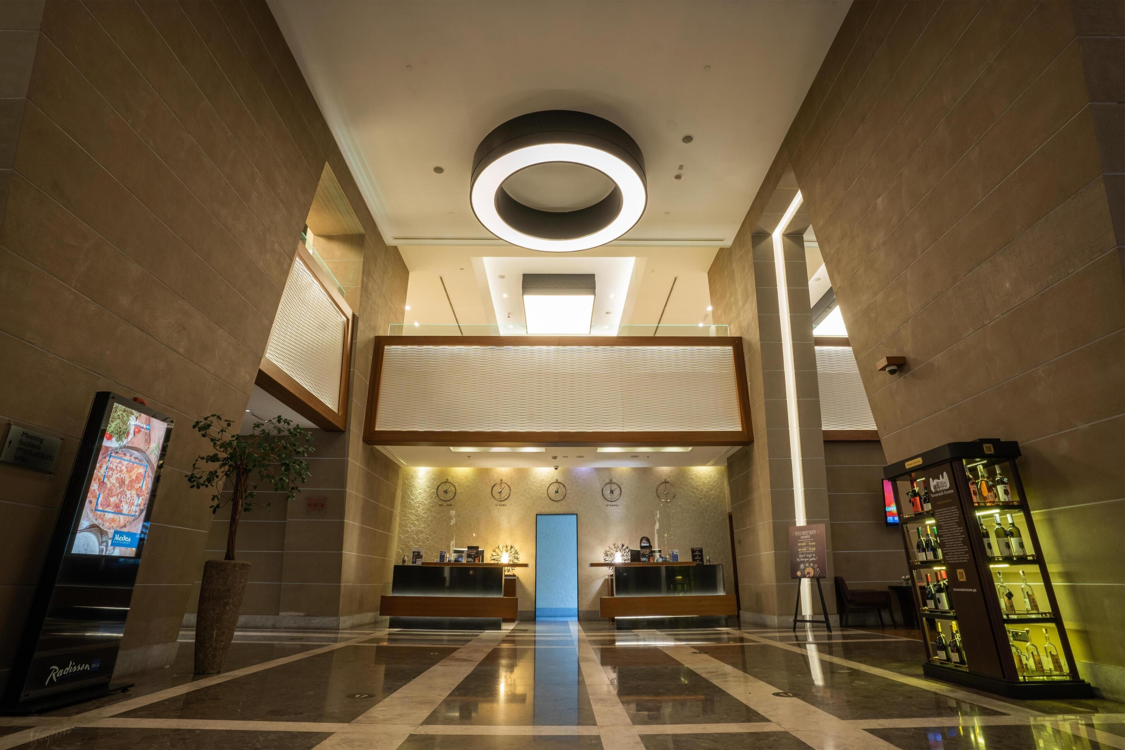 A lobby with a reception desk, a digital screen, a plant, a clock, and a liquor display.