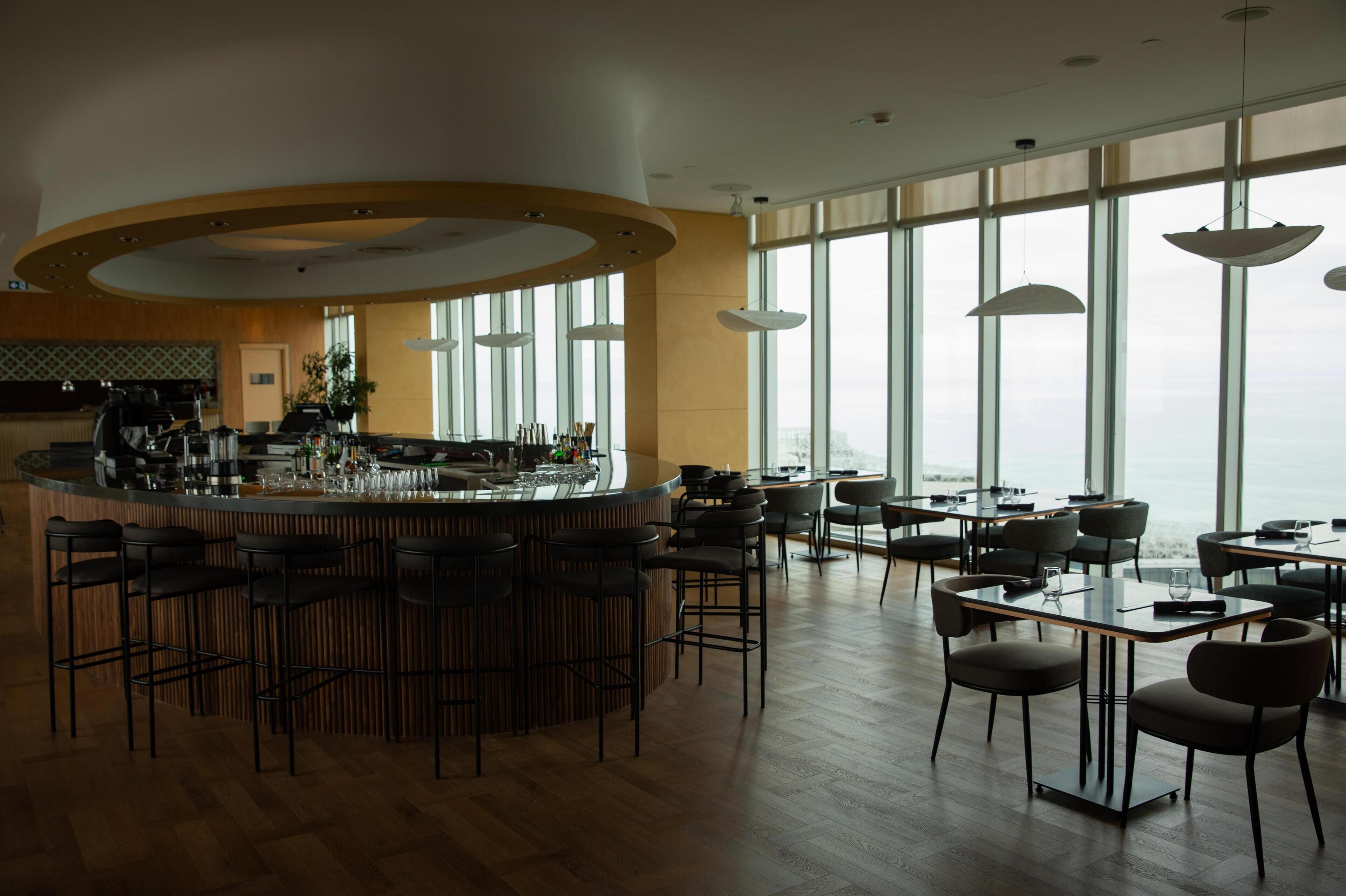 Un área de bar moderna con una barra curva, varios taburetes y un amplio ventanal con vistas a la ciudad.