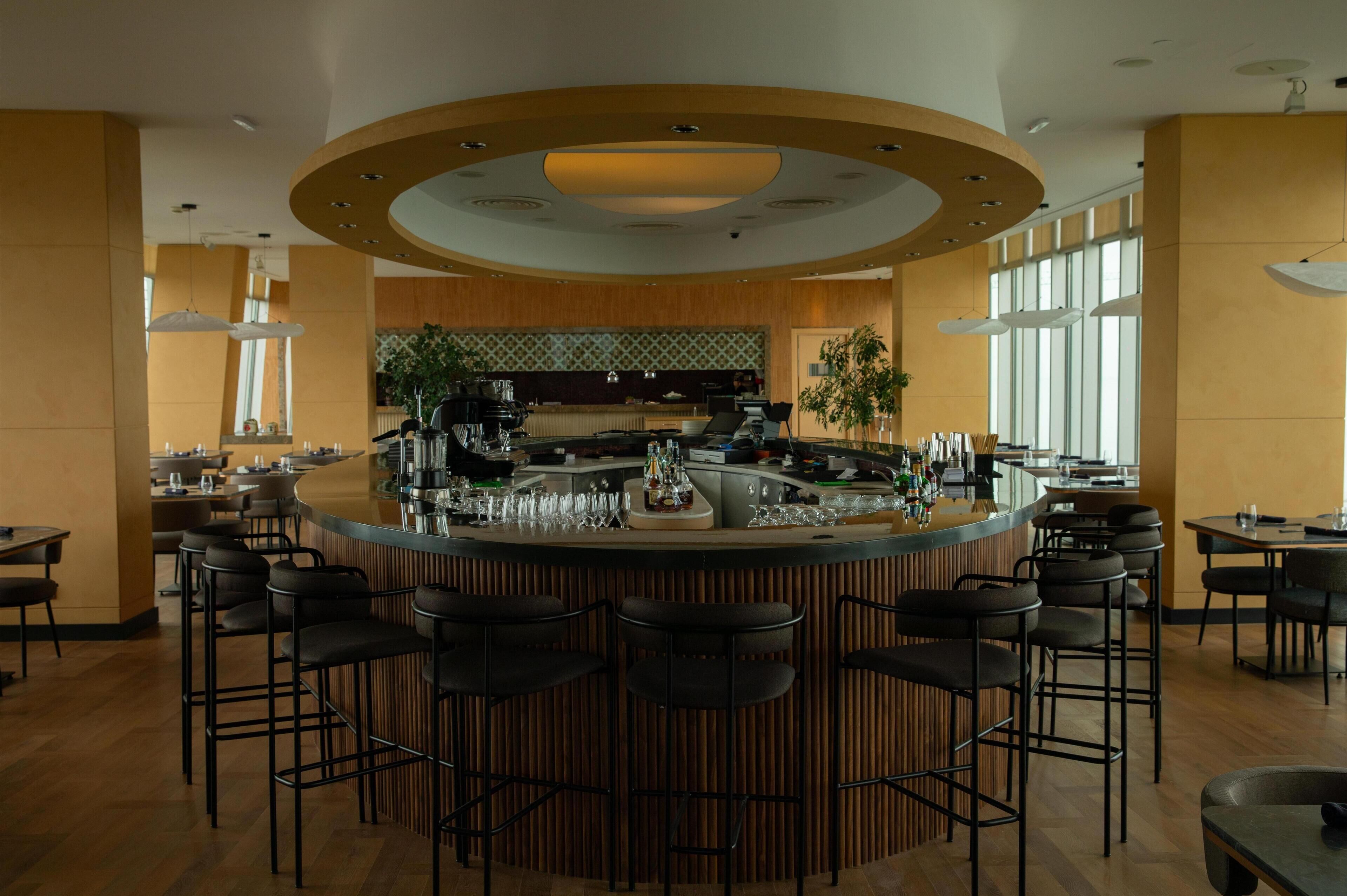 A modern bar area with a curved counter, multiple bar stools, and a kitchen in the background.