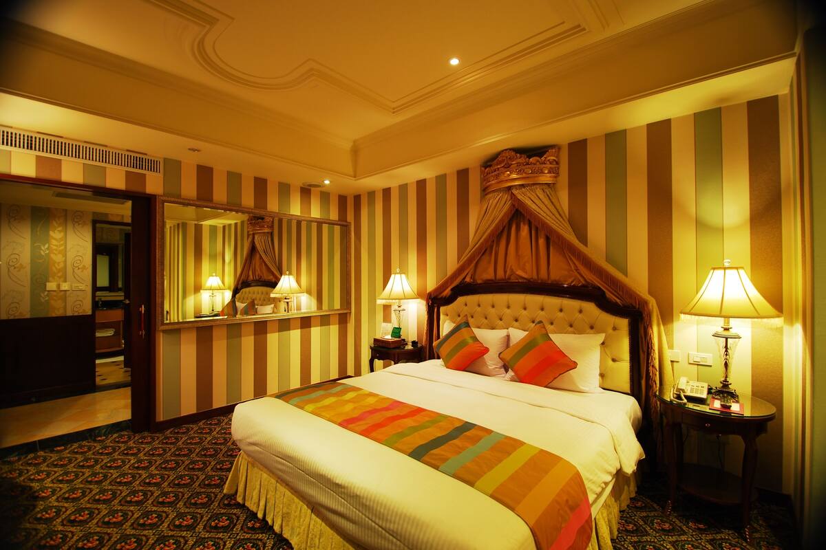 Habitación de hotel con una cama grande, papel tapiz a rayas, dosel sobre la cama, un espejo, lámparas de noche y una alfombra estampada.