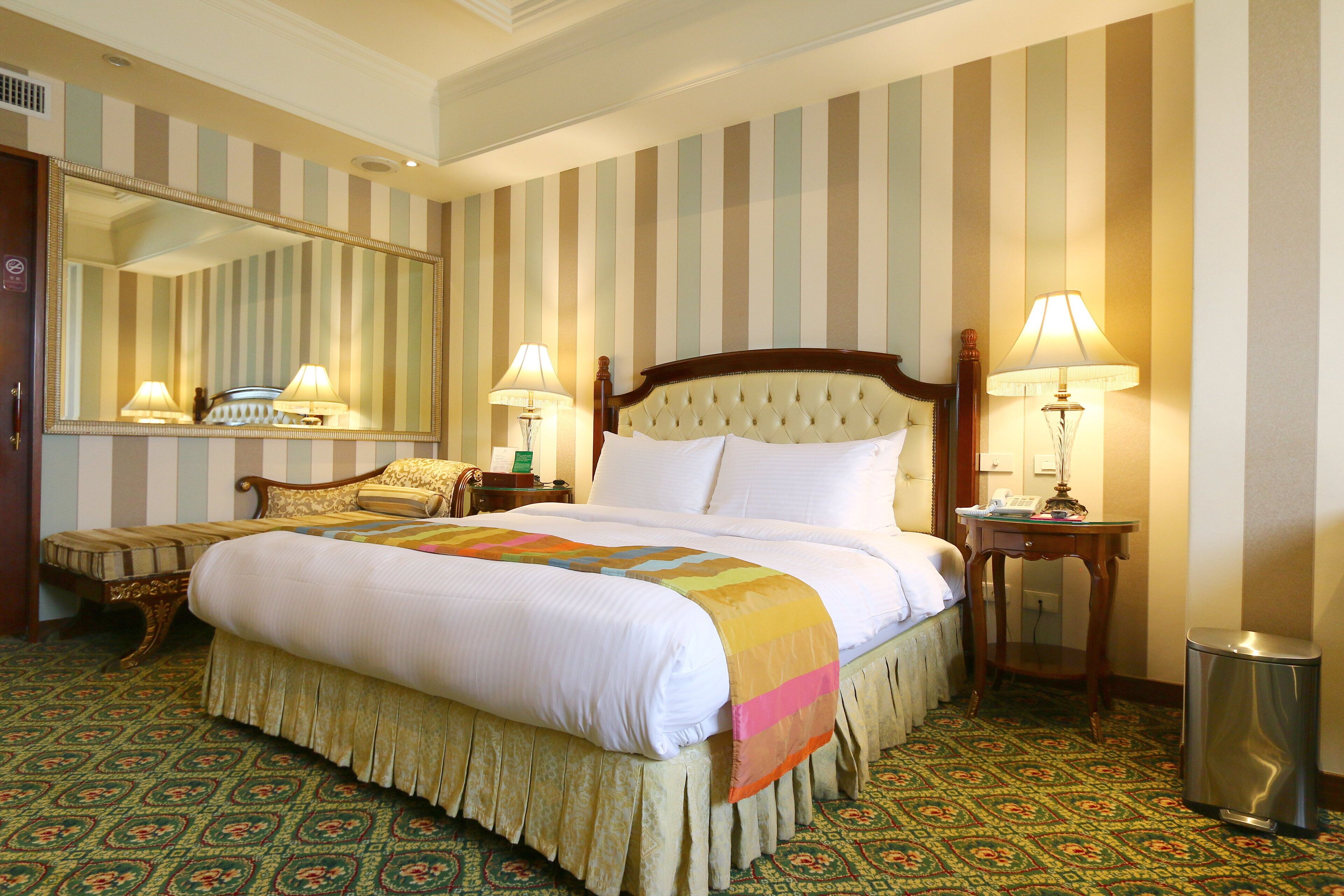 Une chambre d’hôtel avec un grand lit, deux lampes de chevet, un miroir, une petite table et une poubelle.