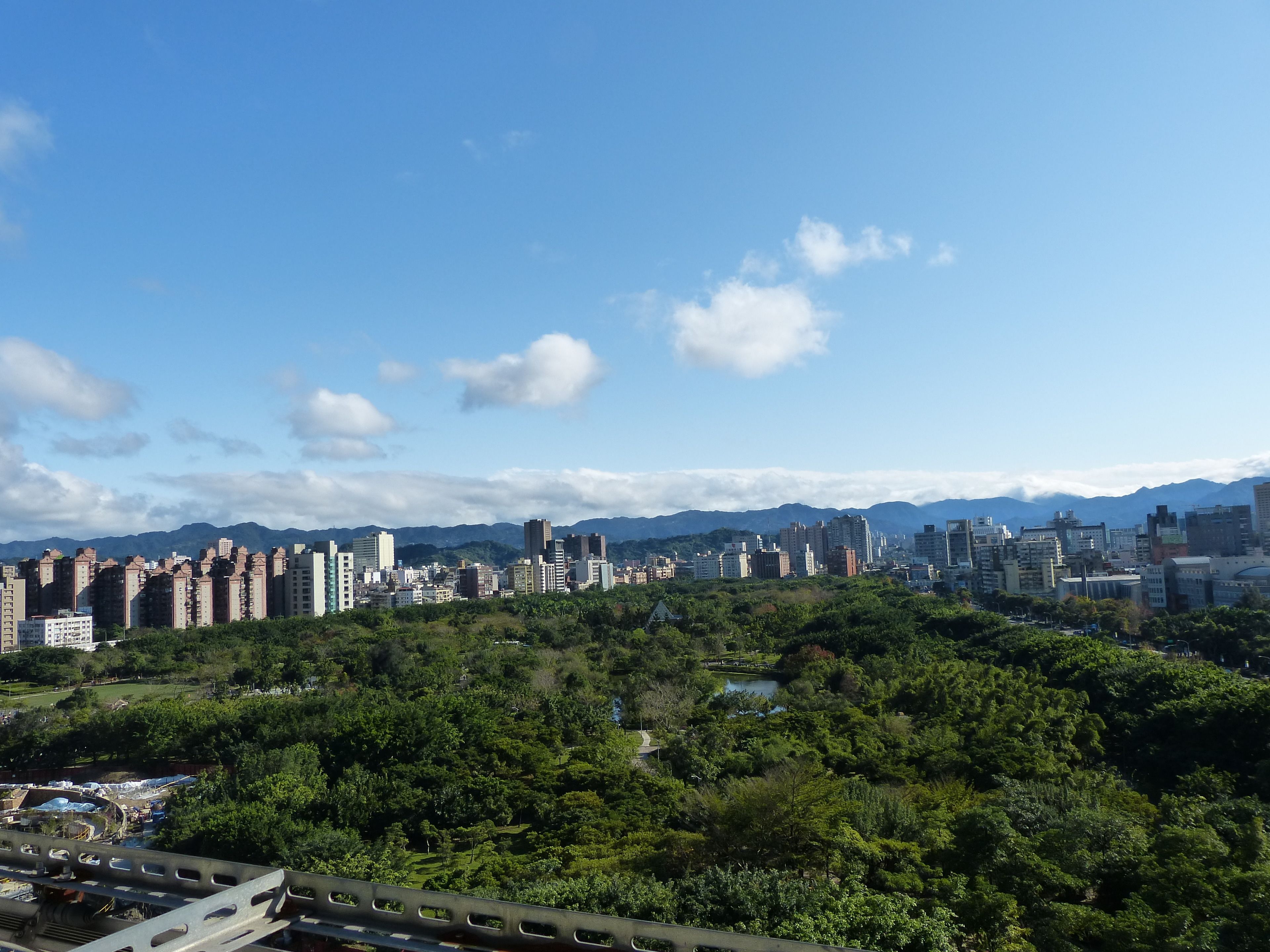 A cityscape with a mix of high-rise buildings and greenery, a clear sky, and mountains in the background.