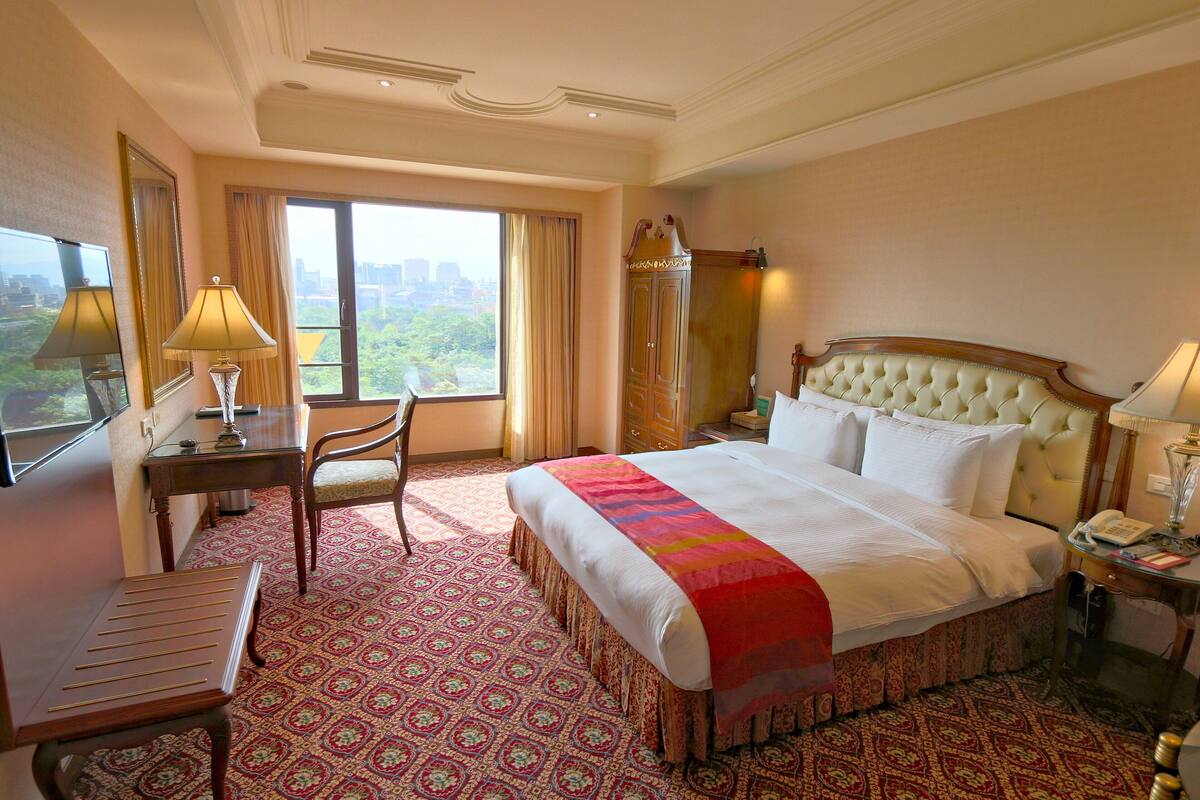 Habitación de hotel con una cama grande, un escritorio, una silla y vistas a un paisaje urbano a través de la ventana.