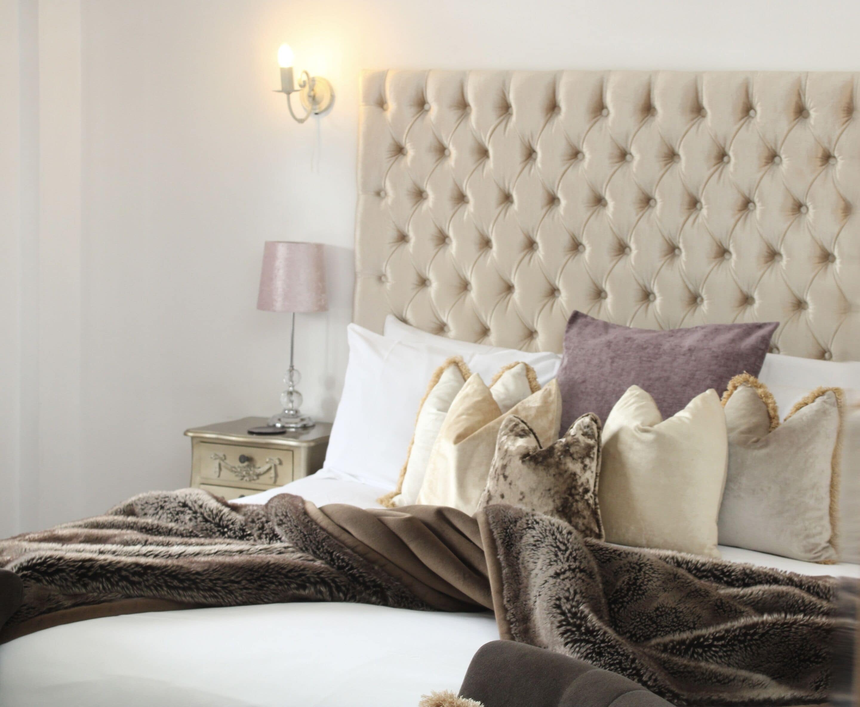 A bedroom with a tufted headboard, a bedside lamp, and a variety of pillows and a blanket.
