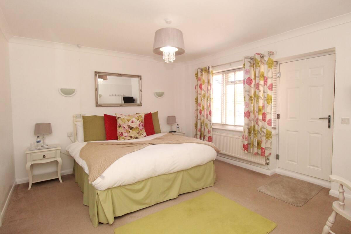 Quarto com cama, mesas de cabeceira, espelho, janela com cortinas florais e porta.