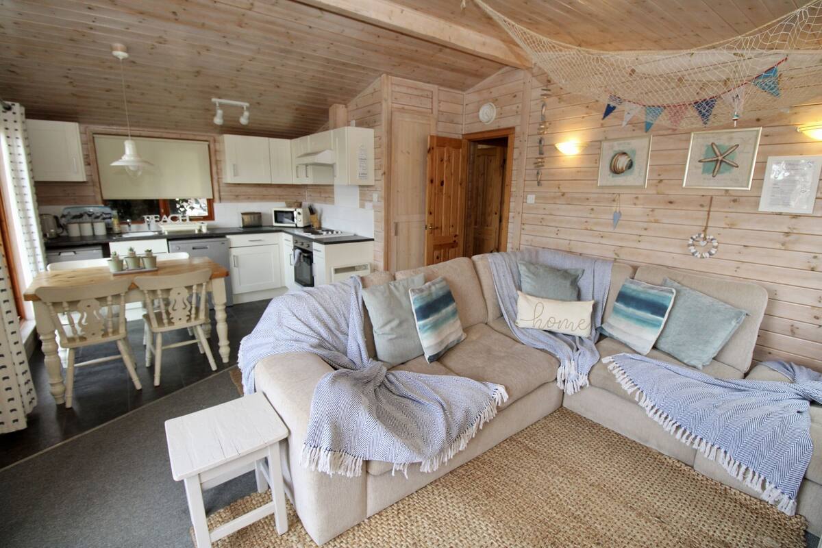 Interior acolhedor de cabana com área de jantar, cozinha compacta e sofá decorado com almofadas.