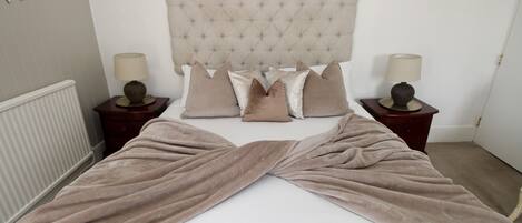 Uma cama bem arrumada com cabeceira bege, duas mesas de cabeceira com abajures e um radiador na parede.