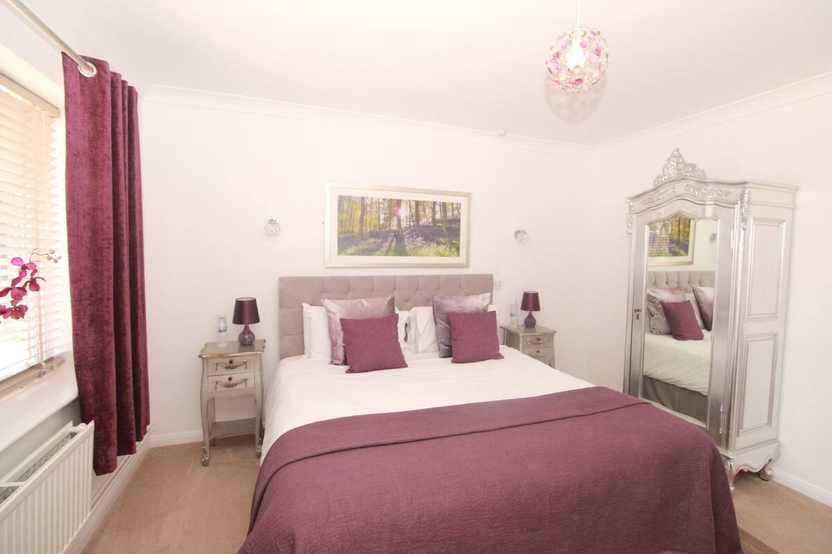 Quarto com cama, mesa de cabeceira, espelho e janela com cortinas.