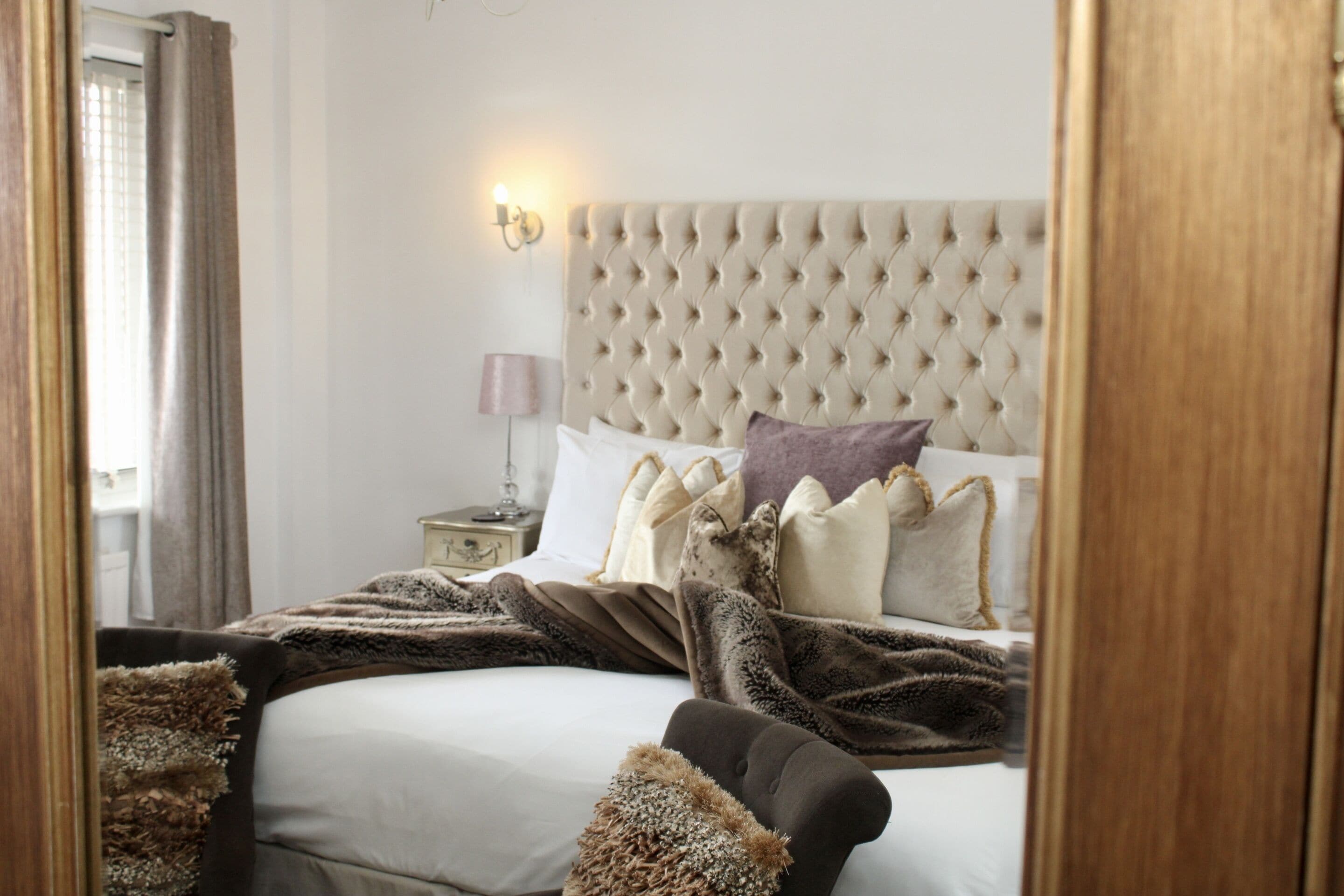 A bedroom with a tufted headboard, a bedside table with a lamp, and a window with curtains.