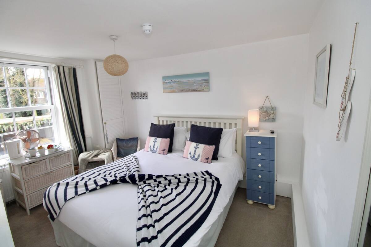 Quarto com cama, mesa de cabeceira, cômoda e janela com cortinas.