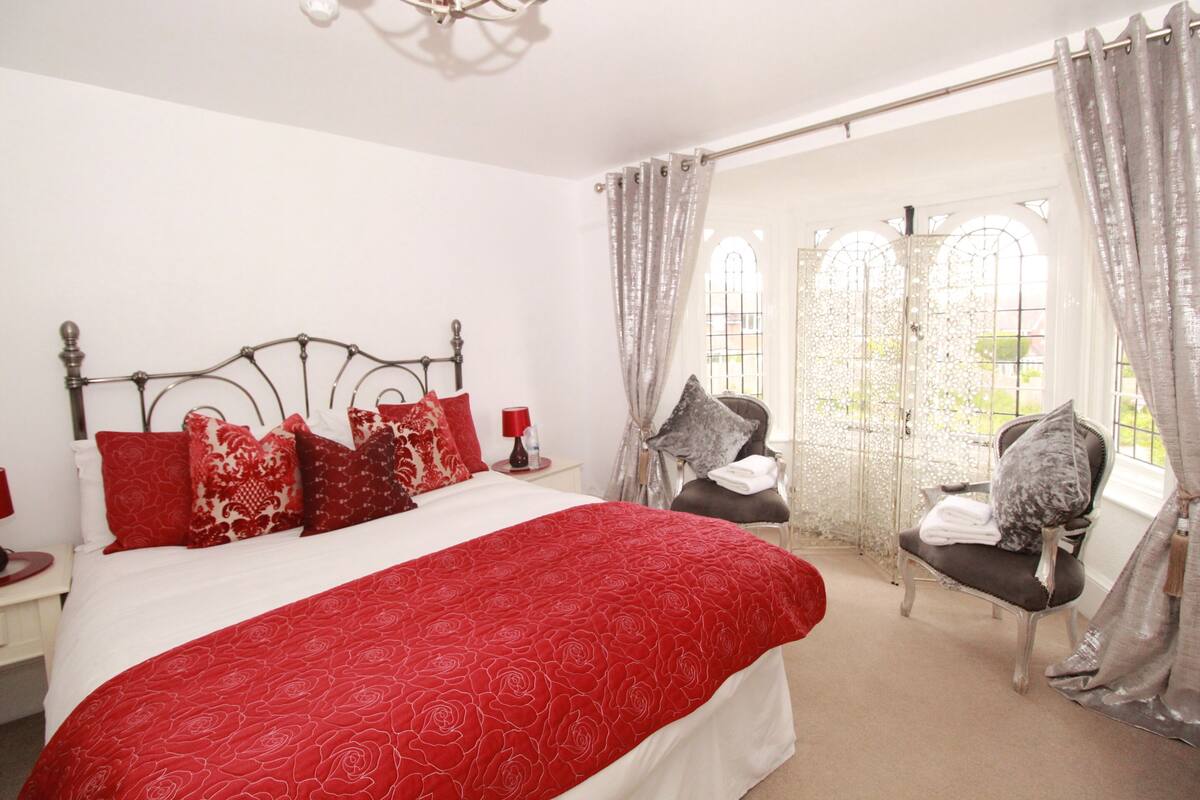 Um quarto com uma cama grande, roupa de cama vermelha e branca, uma mesa de cabeceira com abajur e uma cadeira com encosto decorativo.