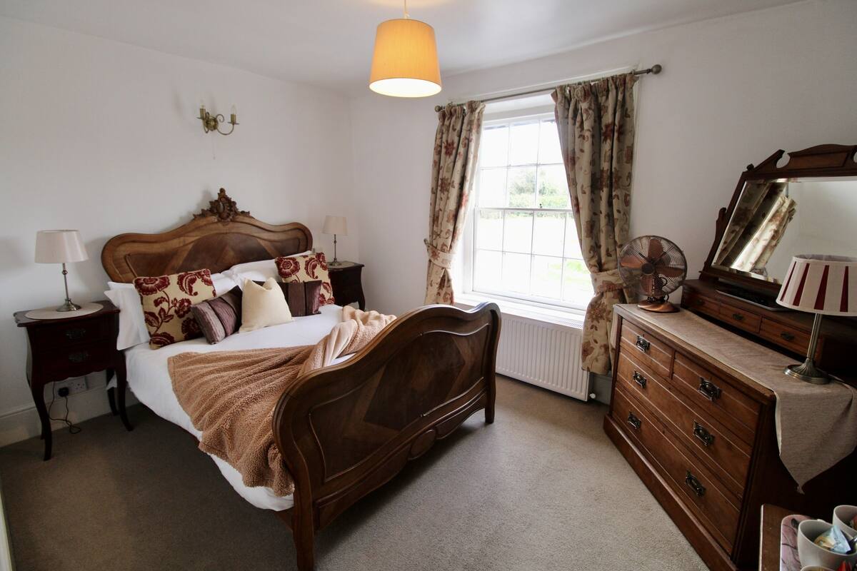 Quarto com cama de madeira, penteadeira, espelho, mesa de cabeceira, abajur e janela com cortinas.