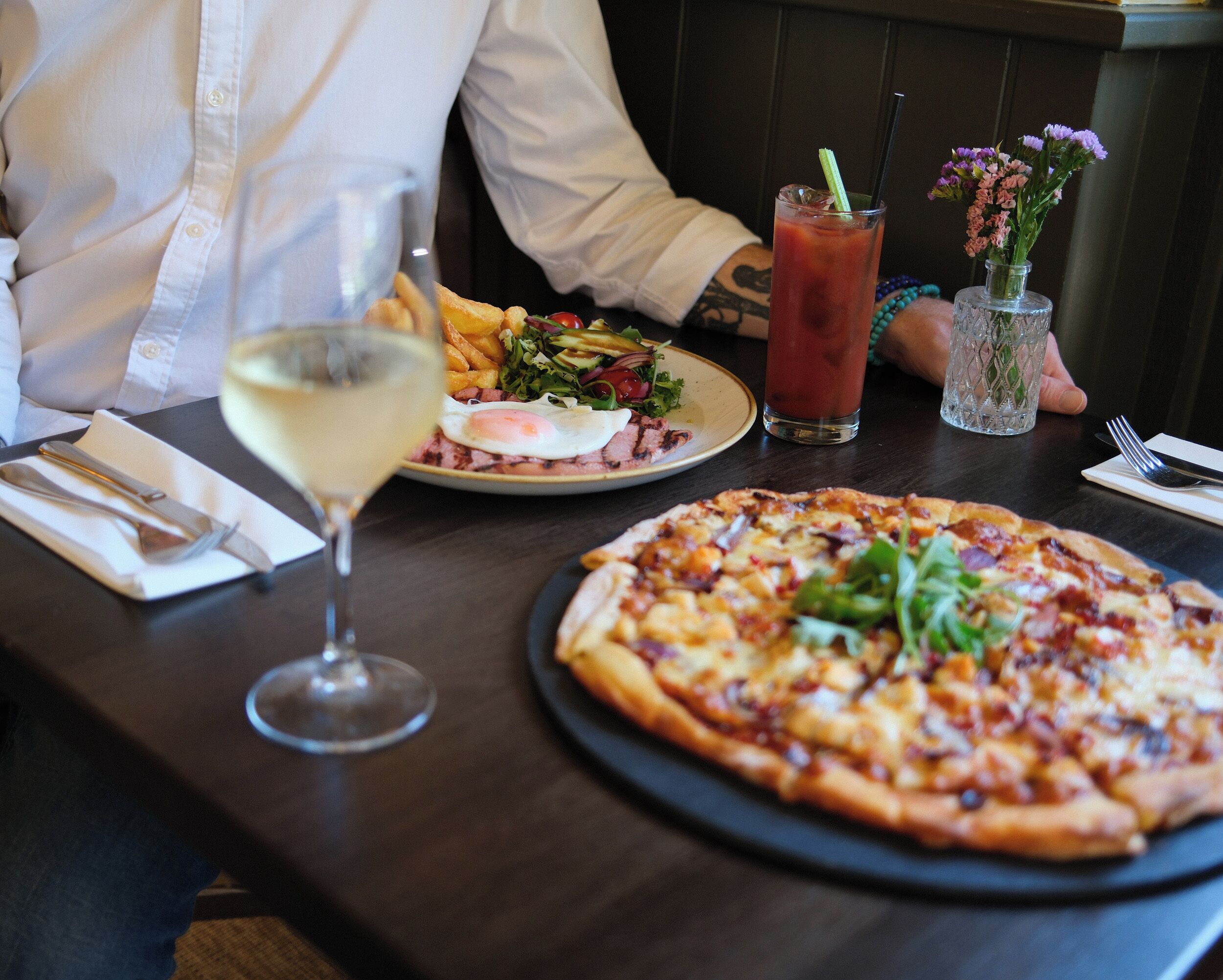 A dining table with a pizza, a salad, a Bloody Mary drink, and a glass of white wine.