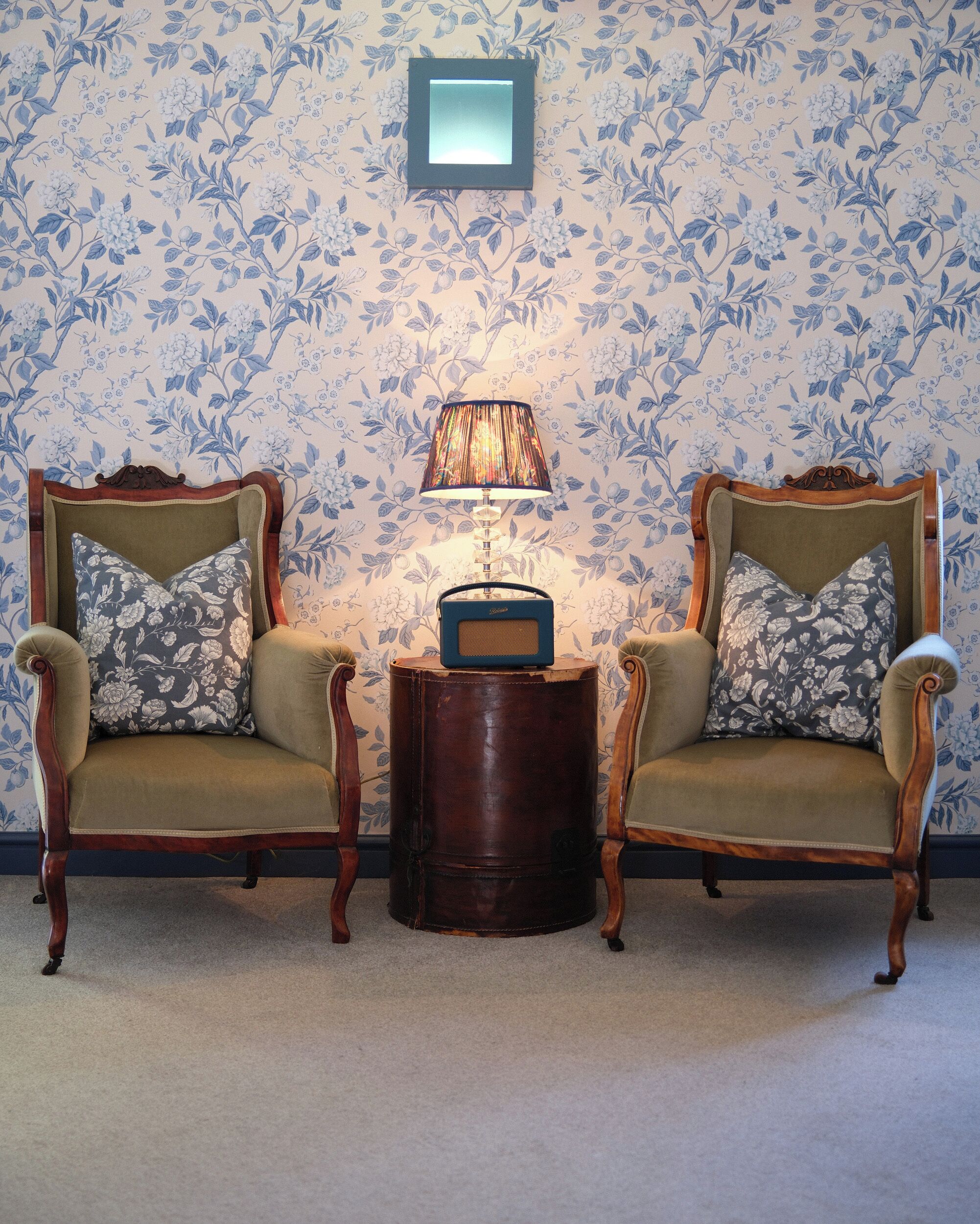 A room with two upholstered chairs with floral cushions, a small wooden side table with a radio, and a wall-mounted picture frame.