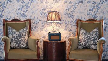 A room with two upholstered chairs with floral cushions, a small wooden side table with a radio, and a wall-mounted picture frame.