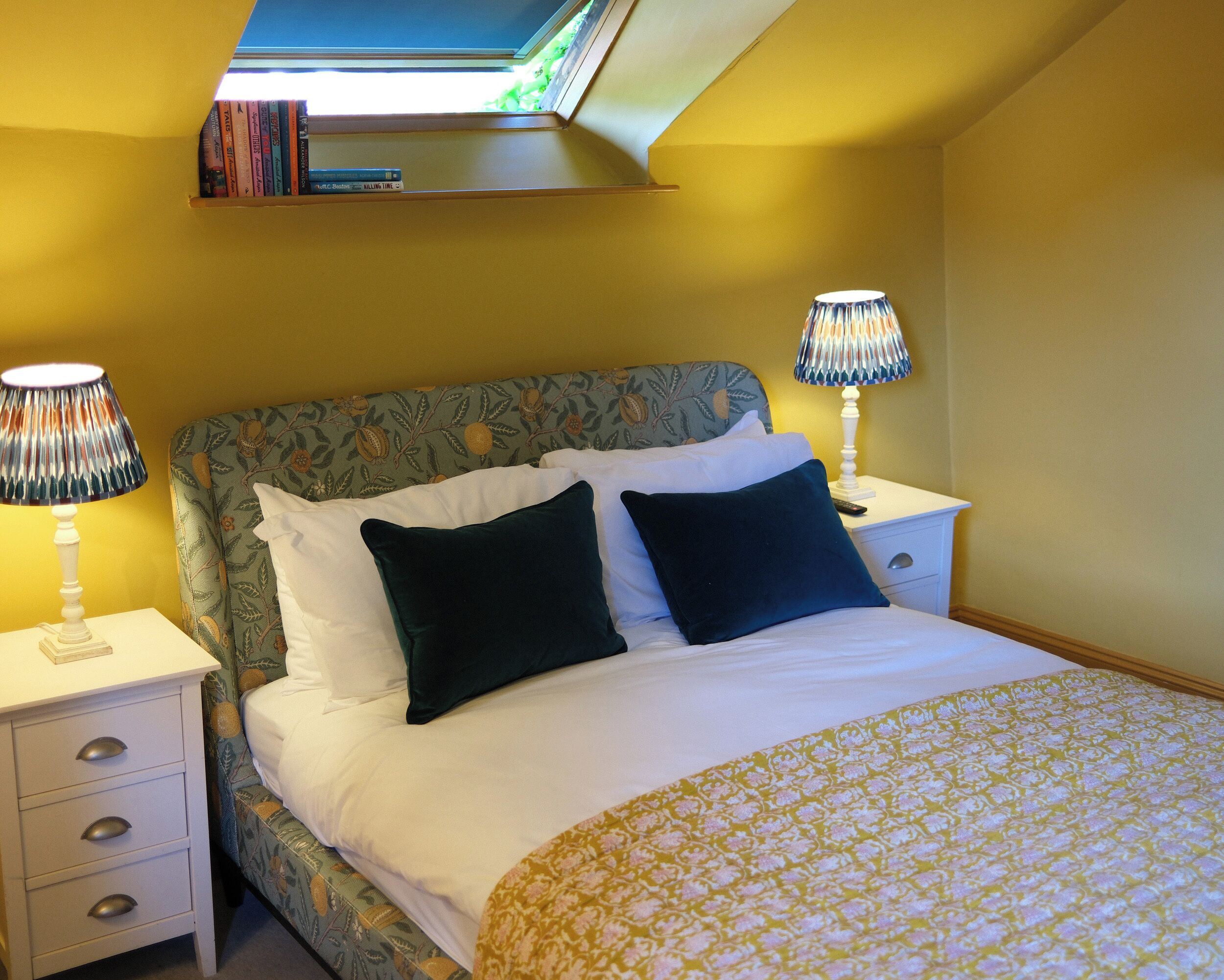 A bedroom with a bed, two bedside tables, a nightstand, a lamp, and a window with books on the shelf.