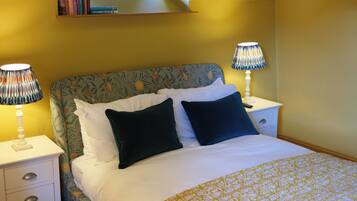 A bedroom with a bed, two bedside tables, a nightstand, a lamp, and a window with books on the shelf.