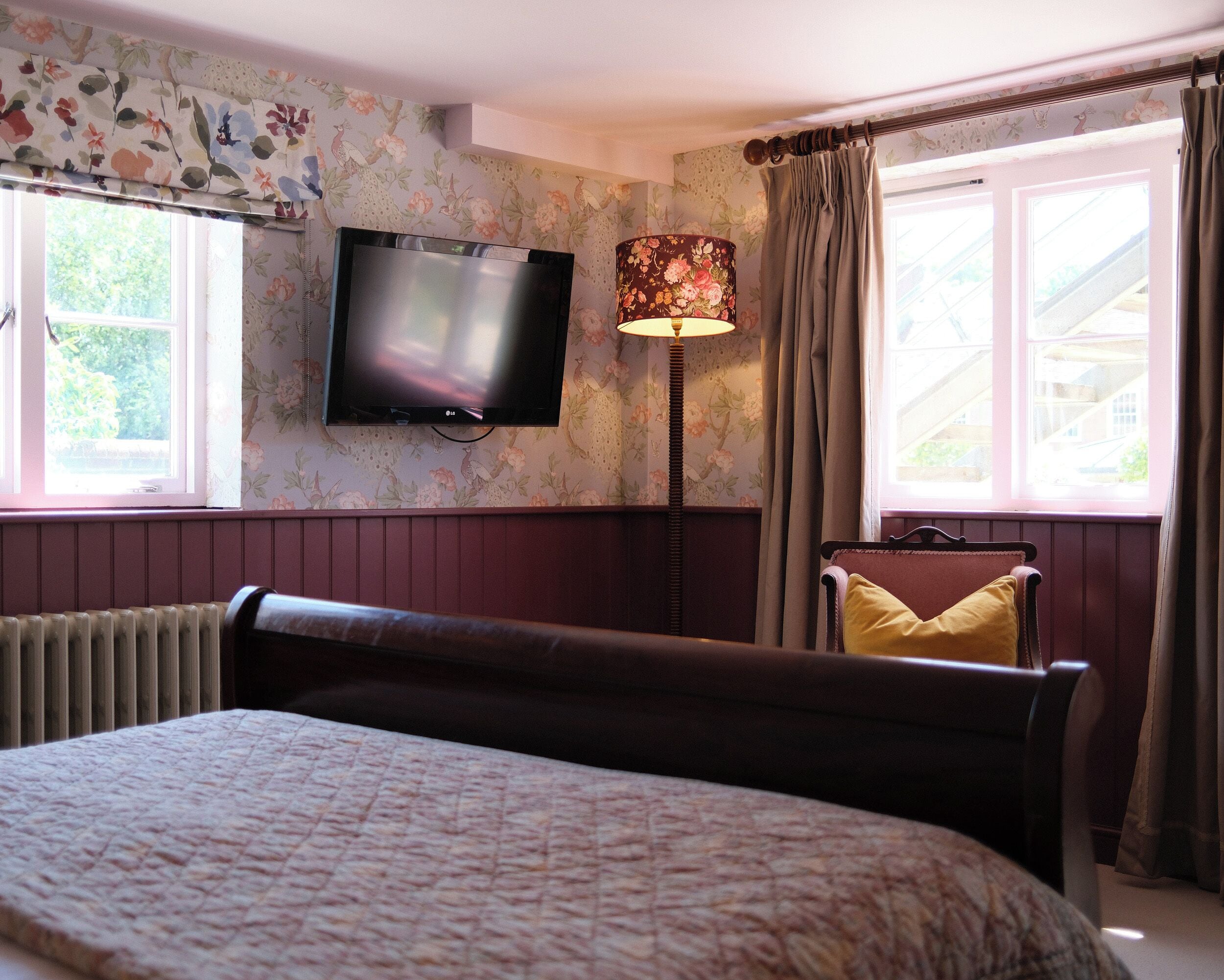 Un dormitorio con una cama, un televisor montado en la pared, una lámpara de pie y una ventana con cortinas.
