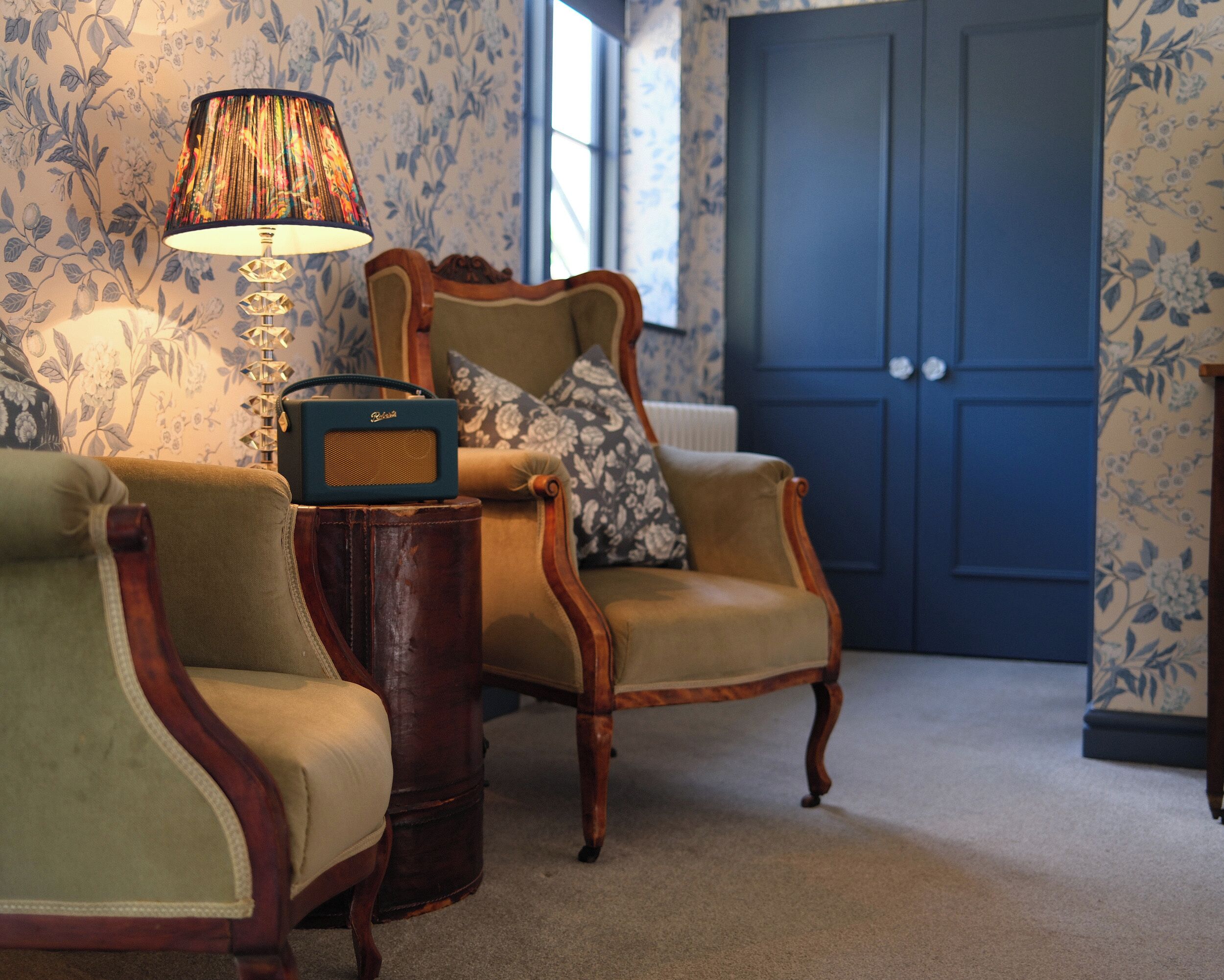 Une pièce avec deux fauteuils, une petite table sur laquelle se trouve une radio, et une lampe. Une porte se trouve au fond de la pièce.