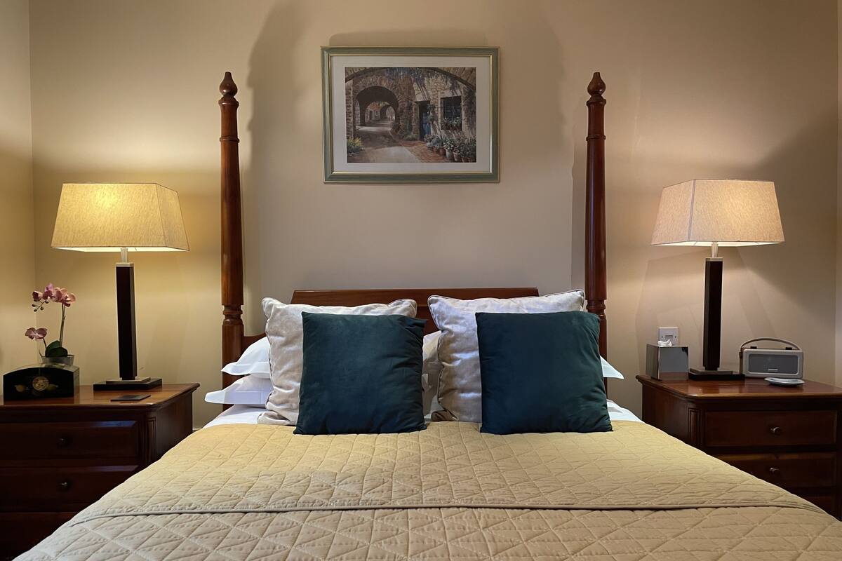 Una cama bien hecha con cubrecama acolchado, dos almohadas, dos lámparas de noche, un cuadro enmarcado en la pared y una mesita de noche con una radio.