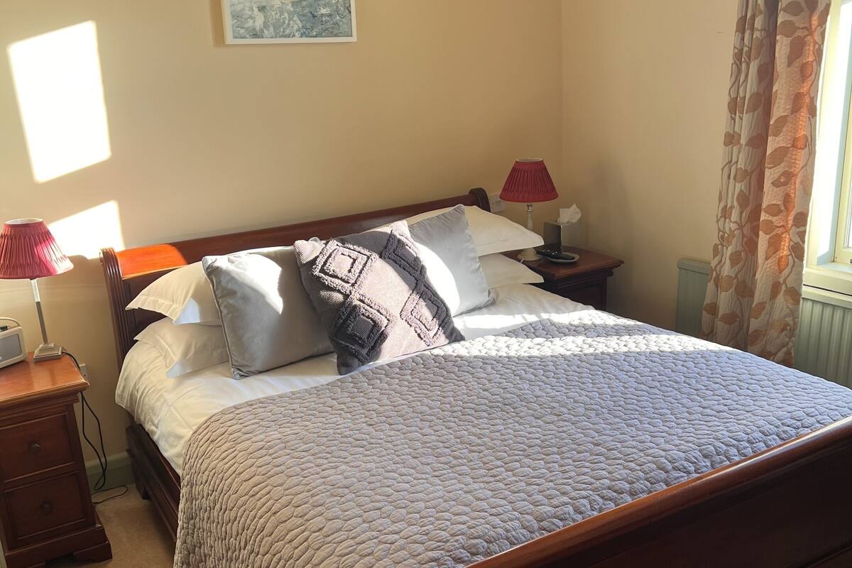 Un dormitorio con cama de madera, una mesita de noche con lámpara, una mesita auxiliar, un cuadro en la pared y una ventana con cortinas.