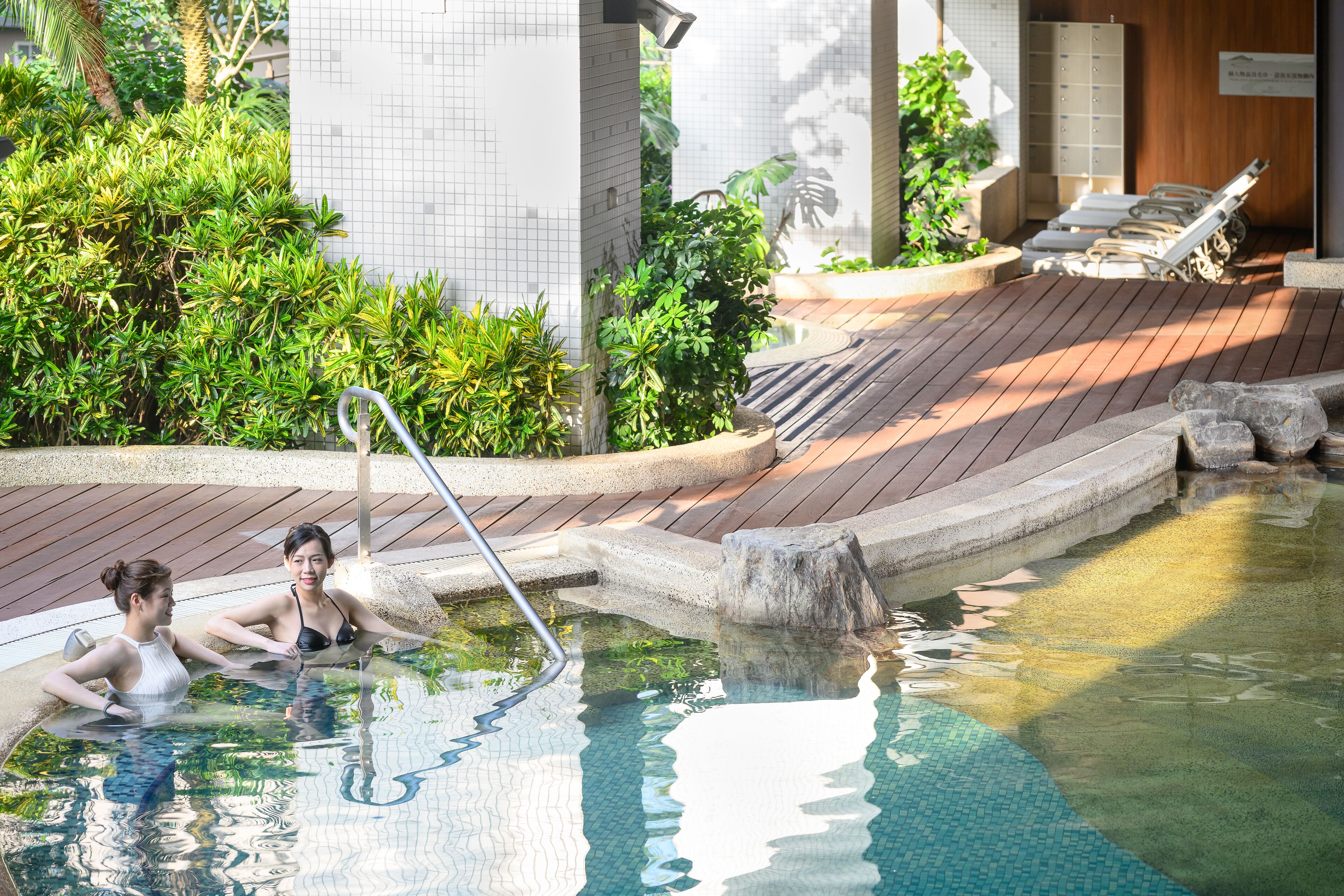 A pool area with two people relaxing, a wooden deck, and a tiled wall in the background.