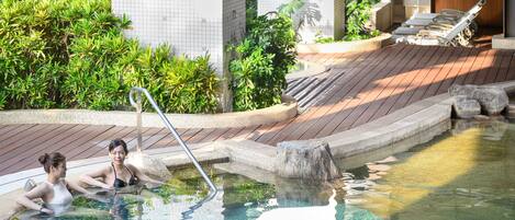 A pool area with two people relaxing, a wooden deck, and a tiled wall in the background.