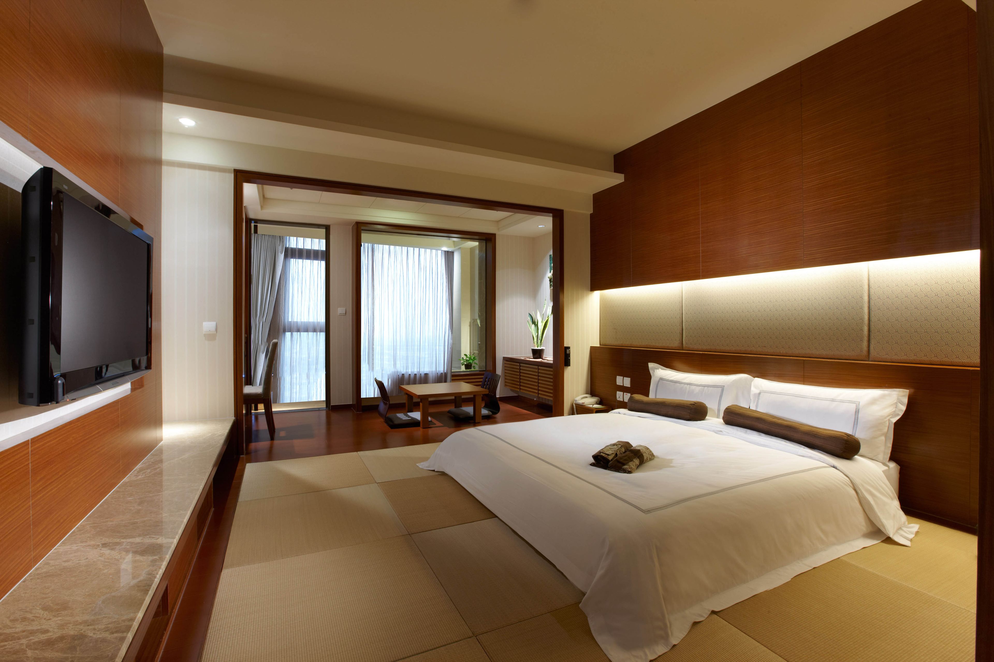 A hotel room with a large bed, a flat-screen TV mounted on the wall, a wooden headboard, and a view of the outside through a glass door.