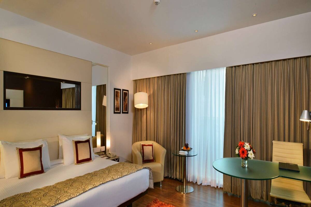A hotel room with a large bed, a desk, a chair, a small table with a vase of flowers, and a large window with curtains.