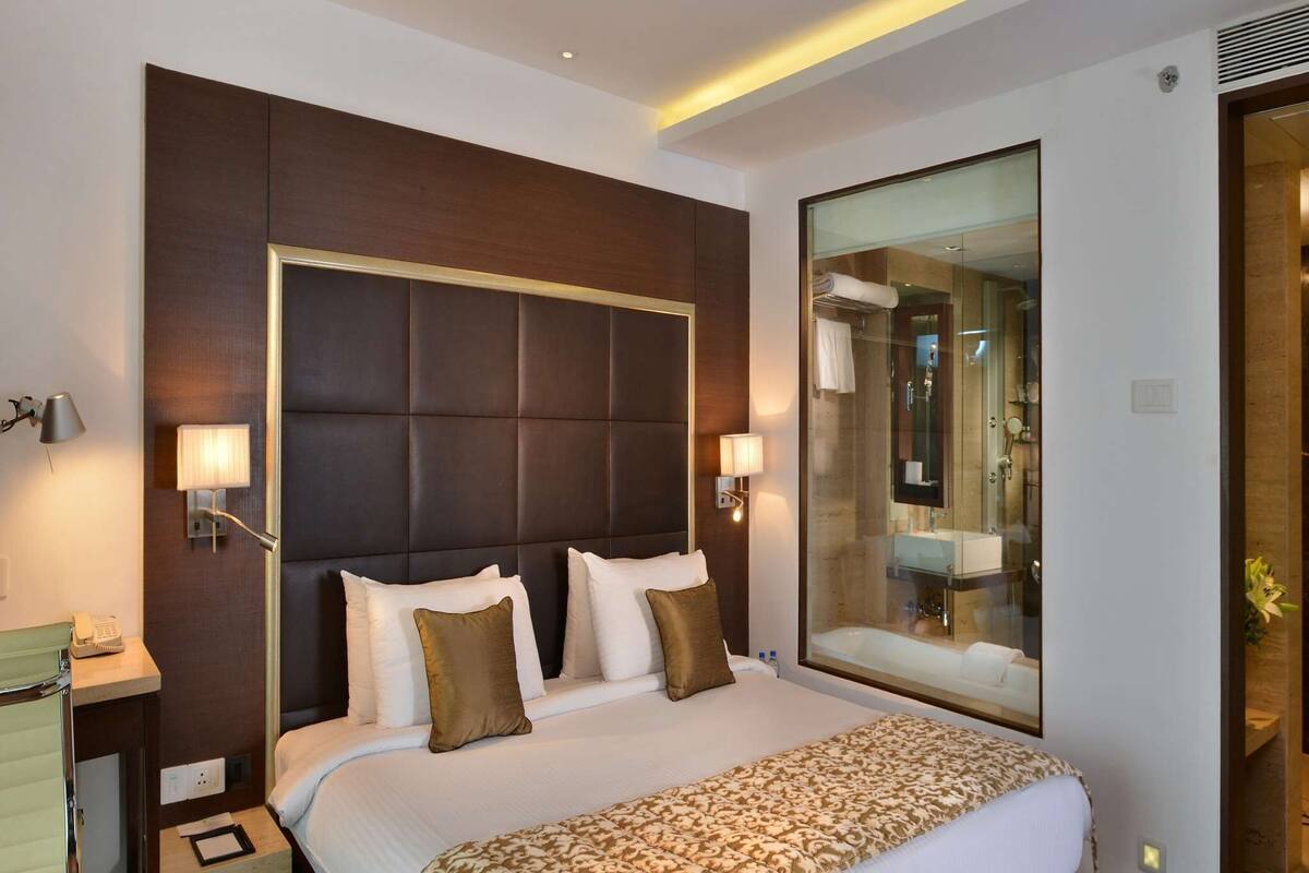 A hotel room with a large bed, a bedside table with a lamp, a mirror, and a view of the bathroom.