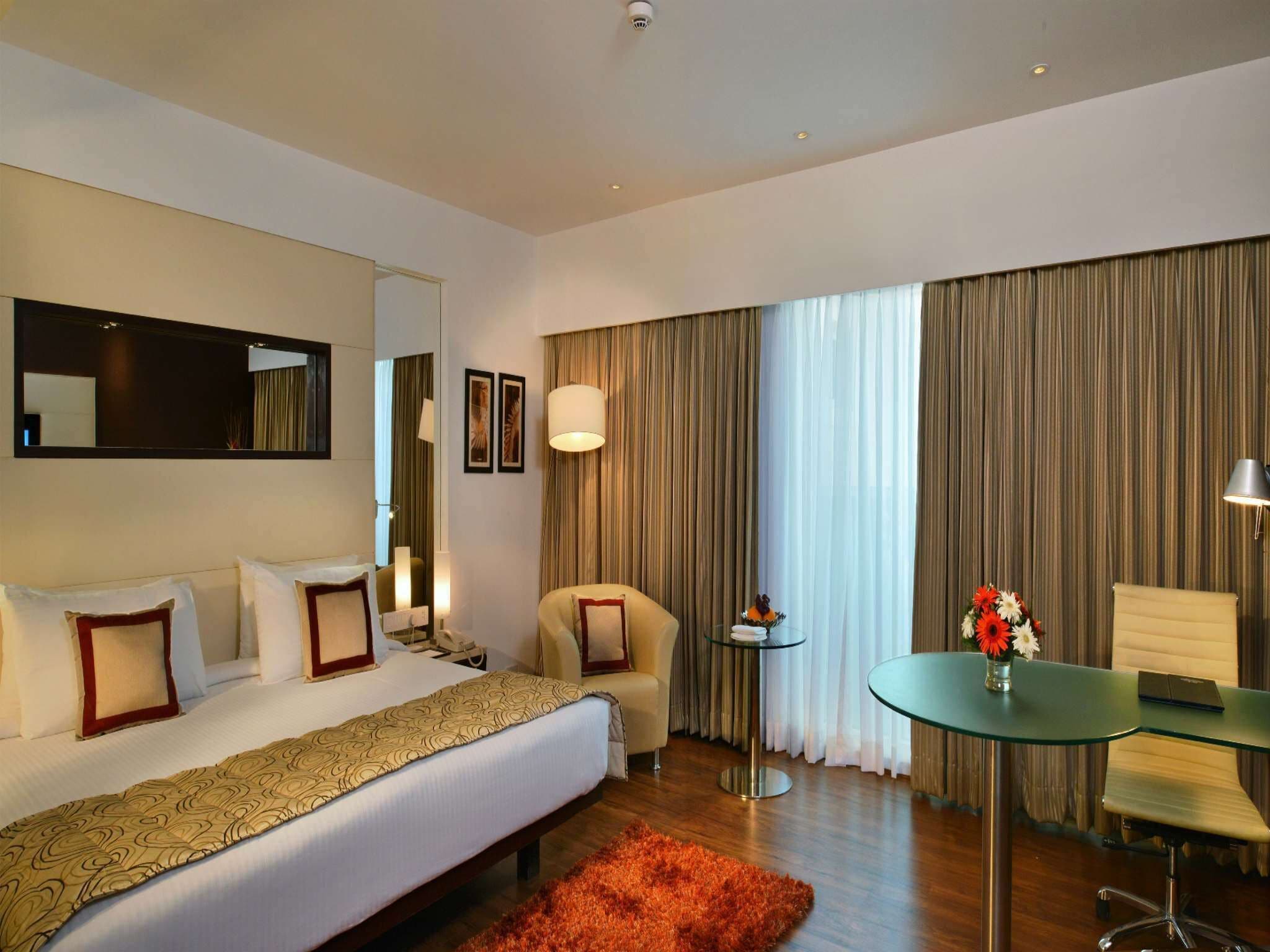 Habitación de hotel con cama, escritorio, silla, espejo y ventana con cortinas.