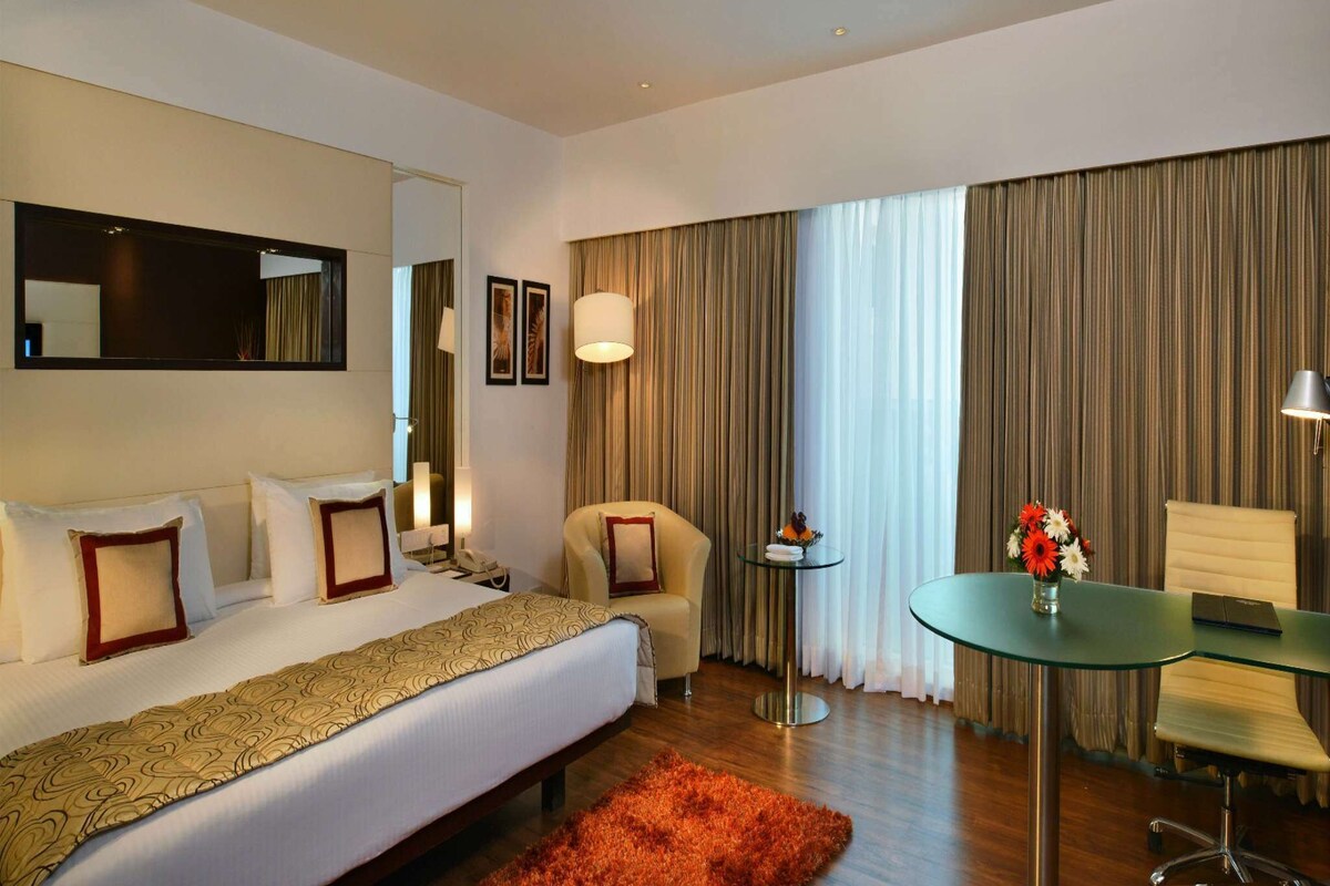 Habitación de hotel con cama, escritorio, silla, espejo y ventana con cortinas.