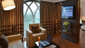 A modern hotel room with a large window, a comfortable sofa, a small armchair, a coffee table with a laptop and a magazine, and a TV displaying a list of destinations. - Majestic Court Sarovar Portico Navi Mumbai (Navi Mumbai)