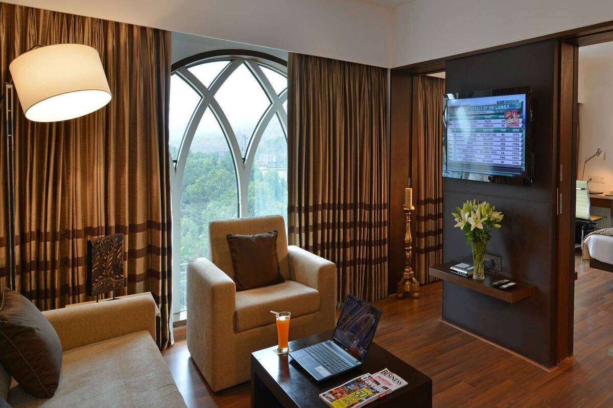 A modern hotel room with a large window, a comfortable sofa, a small armchair, a coffee table with a laptop and a magazine, and a TV displaying a list of destinations.