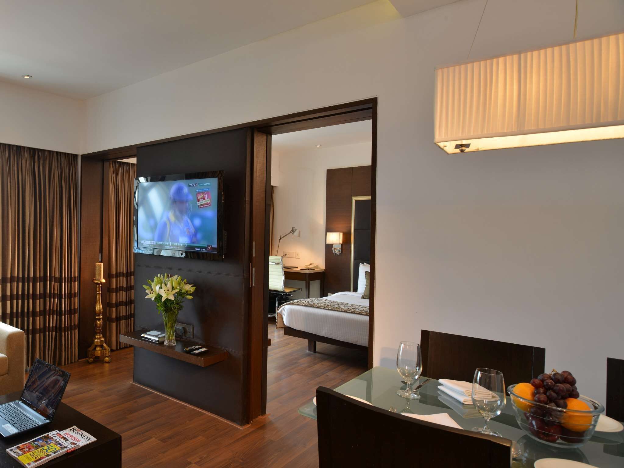 Habitación de hotel con televisión, escritorio con una computadora portátil, mesa de comedor con un bol de fruta y una cama al fondo.