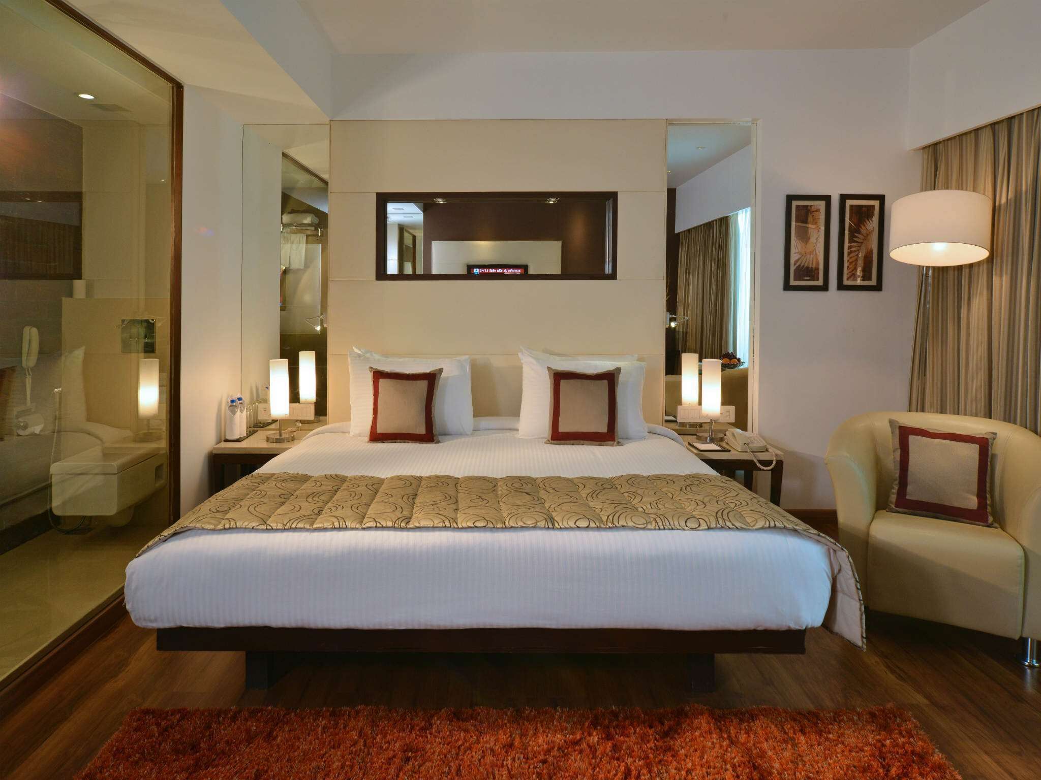 A hotel room with a large bed, a mirror above the bed, a chair, a desk with a lamp, and a red rug.